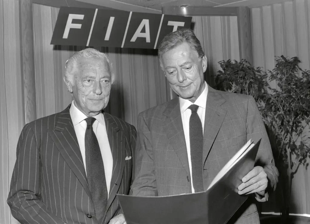 Gianni Agnelli e Umberto Agnelli, em uma foto em preto e branco, revisando um documento sob o logotipo da Fiat.